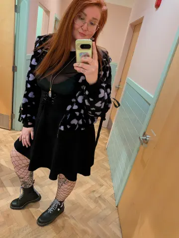 Redheaded woman in black grunge outfit and fishnet tights taking a mirror selfie in a hallway.