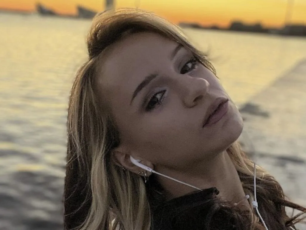 AgataMilk poses outdoors by the water at sunset wearing white earbuds.