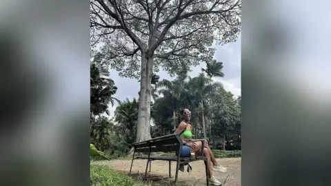 MissyJohansson sits on a park bench wearing a bright green sports bra and blue shorts, smiling under a large, leafy tree.