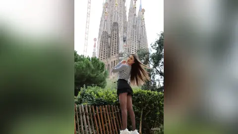 Smiling woman in a striped shirt and black skirt posing in front of the Sagrada Familia cathedral in Barcelona.