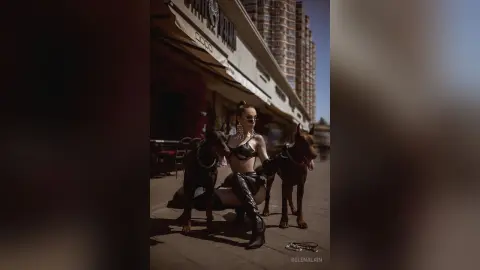Stylish woman in black leather outfit sitting on a city sidewalk with two large dark dogs on a sunny day.