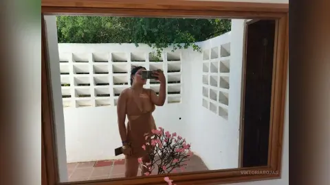 Woman in a tan dress taking a mirror selfie in a bright, white Mediterranean-style patio garden.
