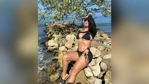 Woman in black bikini and long sleeve crop top posing on rocky coast under sunny summer sky.