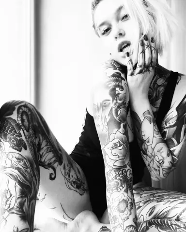 Black and white portrait of a tattooed woman in a black bodysuit sitting in a bright, minimalist setting.