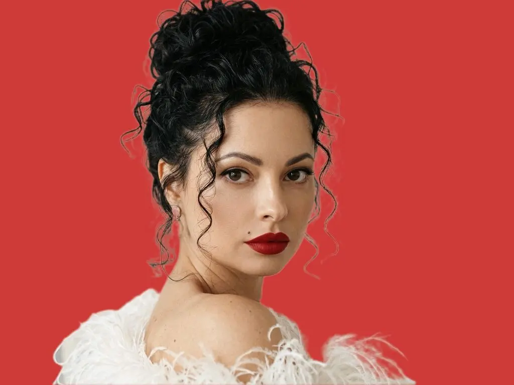Elegant woman with curly dark hair and red lipstick wearing a white feathered outfit against a red background.