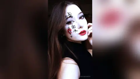 JosefinaHancin poses with long brown hair and red lipstick, featuring black calligraphy tattoos or face paint over her eyes and cheeks.