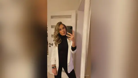 VictoriaWynn poses for a mirror selfie wearing a black turtleneck and a white open cardigan with her long blonde hair down.
