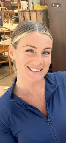 PixiePlayful_ox smiles while wearing a blue zip-up top, showing her blonde hair and nose piercing in an outdoor cafe setting.