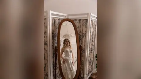 Elegant woman in a white lace wedding dress posing in front of a vintage oval mirror in a bright room.