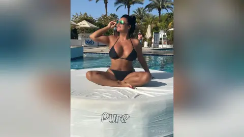 MissyJolie sits cross-legged on a white cushion by a swimming pool wearing a black bikini and sunglasses.
