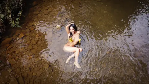 Smiling brunette woman in a floral bikini sitting in a shallow rocky stream surrounded by nature.