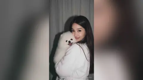 Smiling woman in a white sweater hugging a fluffy white Pomeranian dog in a cozy indoor setting.