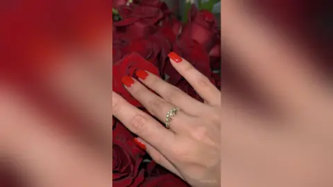 Close-up of a woman's hand with red nails wearing a gold ring against a romantic background of red roses.