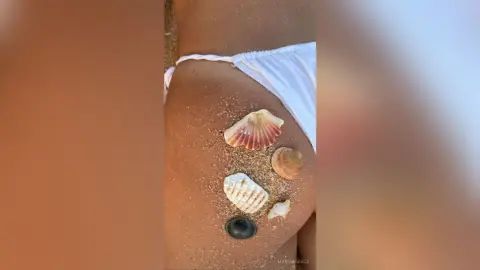 MarisaGrace shows her bare stomach and lower belly, decorated with several seashells on a sandy beach while wearing a white bikini bottom.