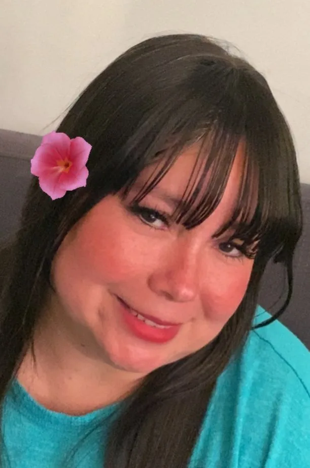 Smiling woman with a pink flower in her hair wearing a teal shirt in a cozy indoor setting.