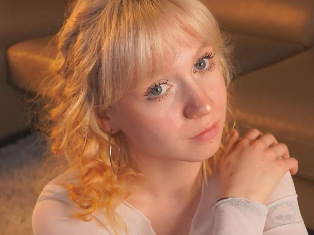 Blonde woman with wavy hair in a white top looking thoughtfully at the camera in a cozy indoor setting.