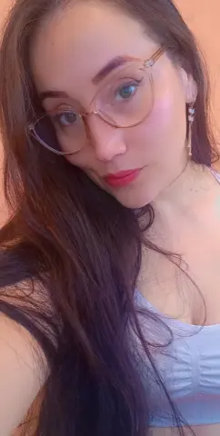 Smiling woman with long dark hair and gold glasses wearing a white tank top in soft indoor lighting.