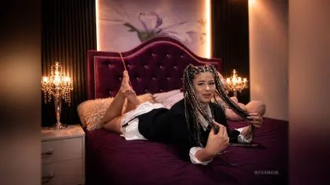 Smiling woman with long braids relaxing on a purple velvet bed in a cozy, moody bedroom setting.