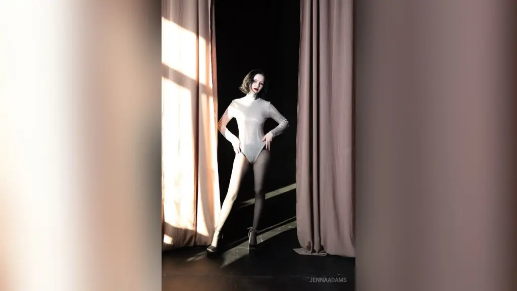 Woman with neck tattoos wearing a red velvet dress posing against a white wall in moody lighting.