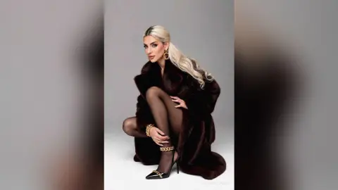 JessycaPage poses in a dark brown fur coat and black sheer tights, looking at the camera with long blonde hair.