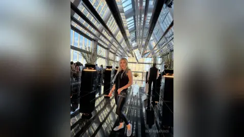 Stylish woman in a black sleeveless top and leather pants standing in a modern glass atrium with reflective floors.