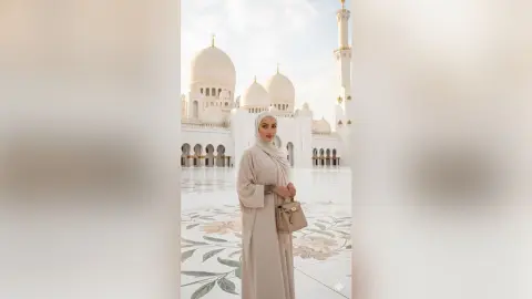 Elegant woman in a beige modest abaya standing before the white Sheikh Zayed Grand Mosque at sunset.