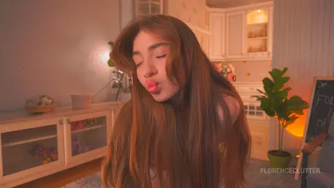 Young woman with long brown hair making a playful kissy face in a cozy, warm-lit bedroom setting.
