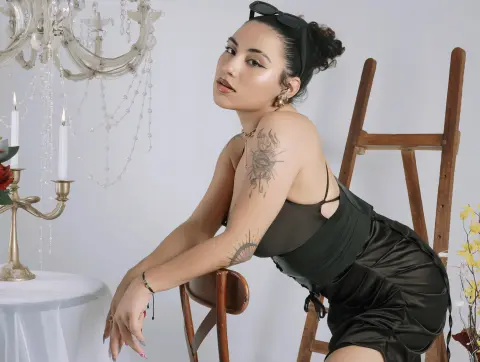 BrianaKnox — Tattooed woman in a black satin outfit sitting in a bright studio with a vintage chandelier and wooden ladder.