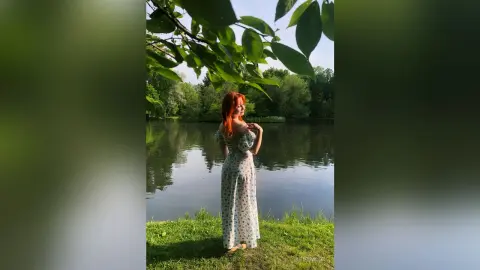 Redheaded woman in a white floral maxi dress standing by a peaceful lake in a lush green forest setting.