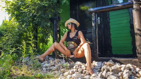 MissyJolie sits outdoors on a dark wooden porch wearing a black tank top and a straw hat, surrounded by rocks and green foliage.