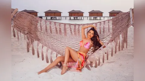 Carine relaxes in a woven hammock on a white sand beach, wearing a pink patterned bikini and a matching sarong.