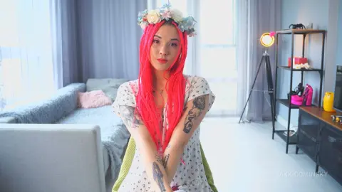 VikkiCominsky poses in a white patterned top and a floral headpiece, showing off her long red hair and arm tattoos.