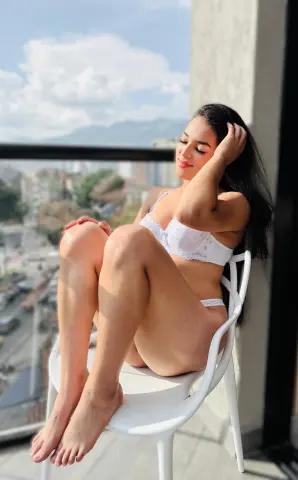 Smiling young woman in white lace lingerie relaxing on a balcony overlooking a sunny city skyline.