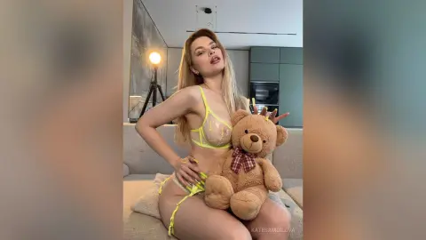 Blonde woman in neon yellow lingerie sitting on a sofa holding a brown teddy bear in a modern living room.