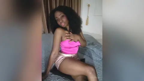Young woman in a pink strapless top and satin skirt sitting on a bed in a bright bedroom.
