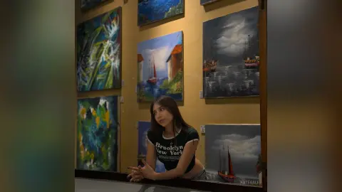 KateGale poses in a black t-shirt with white text, leaning against a table in front of several colorful paintings in a room with tan walls.