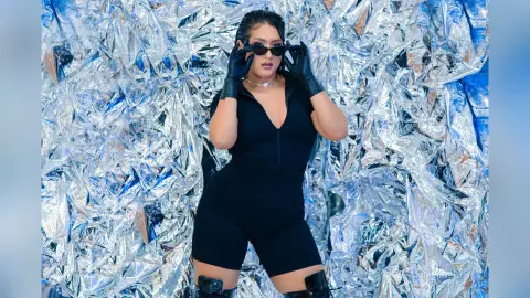 KimRoberth poses in a black sleeveless bodysuit and black sunglasses against a shiny silver foil background.