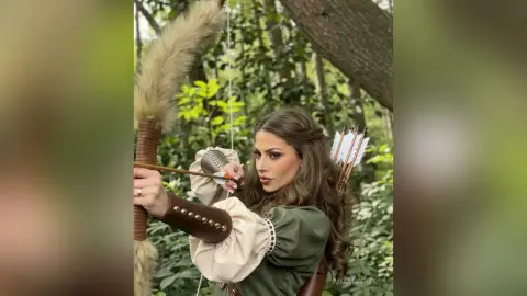 AnaMathers holds a wooden bow and arrow, wearing a long-sleeved green top and brown leather arm guard in a forest setting.