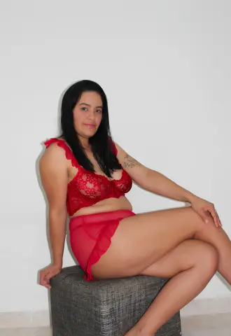 Smiling woman in red lace lingerie sitting on a grey ottoman against a white wall background.