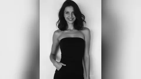 Smiling young woman in a black strapless dress posing against a plain wall in a black and white portrait.