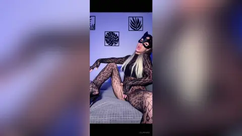 Woman in black lace bodysuit and mask reclining on a bed in a modern room with leaf wall art.
