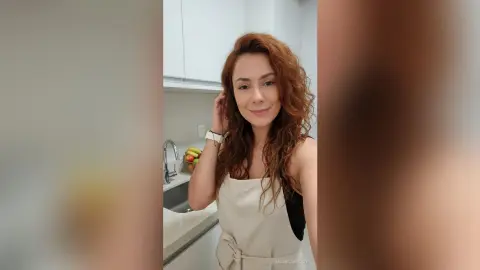 Smiling woman with wavy red hair wearing a cream sleeveless top in a bright modern kitchen.