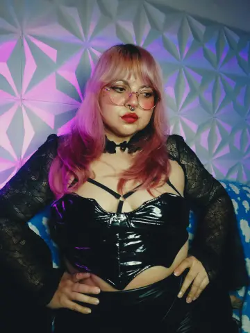 Woman with pink hair in black lace outfit and glasses posing against a geometric textured wall.