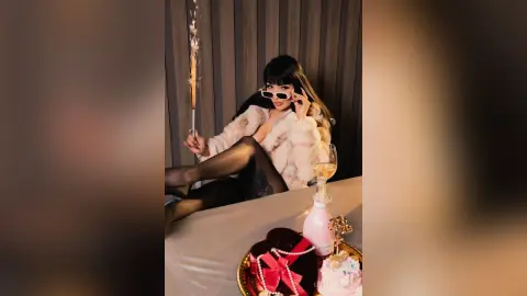 Glamorous woman in white faux fur and sunglasses holding a sparkler next to champagne and cake at a party.