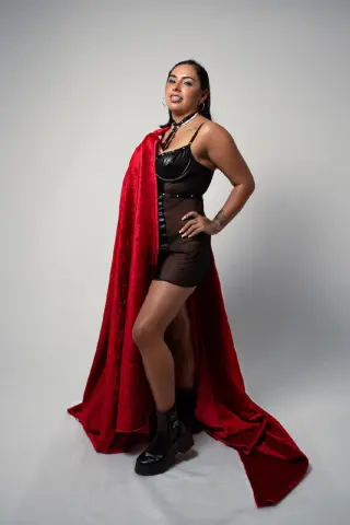 Confident woman in a black bodysuit and long red velvet cape posing against a white studio background.