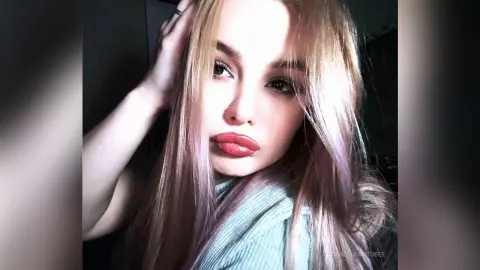 Blonde woman with long hair and red lipstick posing in a light blue sweater for a soft, glamorous selfie.