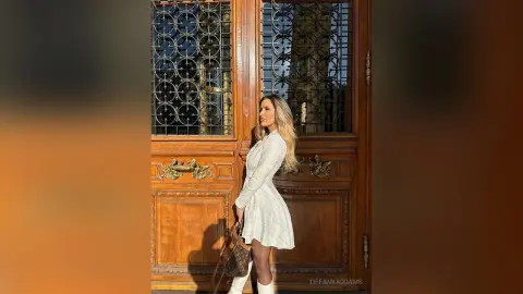 TiffanyAddams poses in a white plaid dress and white boots, standing in front of a large, ornate wooden door with metal decorative grilles.
