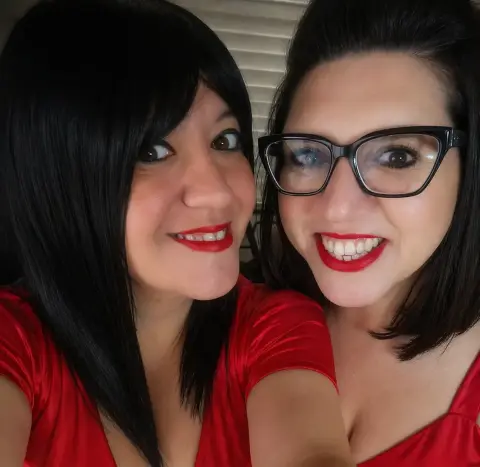 Sinfulmarkedqueens and a friend with dark hair and red lipstick smile closely together in red tops.