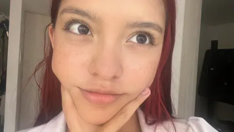 AlyMartini looks into the camera with a playful smirk, resting her chin on her hand with her red hair framing her face.