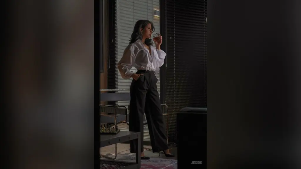 Jessie — Elegant woman in white blouse and black trousers holding a drink in a moody, dimly lit modern interior.